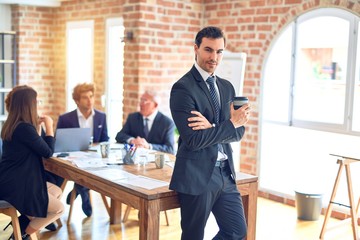 Group of business workers smiling happy and confident working together in a meeting. One of them, standing with smile on face looking at camera drinking coffee at the office.