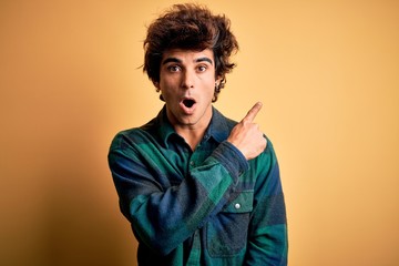 Young handsome man wearing casual shirt standing over isolated yellow background Surprised pointing...