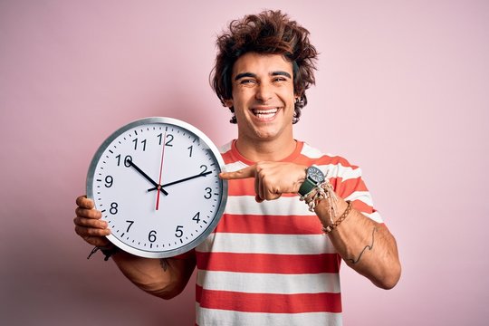Young handsome man holding big clock standing over isolated pink background very happy pointing with hand and finger