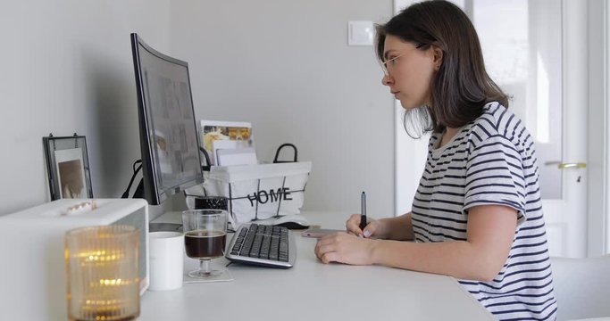 Medium shot of Young hipster girl Working on Digital Drawing Tablet at His Desktop Computer. Distance Work While Coronavirus COVID-19 Pandemic. Her Workplace is Covered with Sunlight.