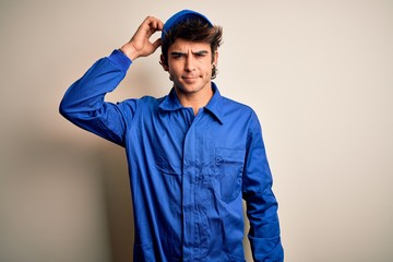 Young mechanic man wearing blue cap and uniform standing over isolated white background confuse and wonder about question. Uncertain with doubt, thinking with hand on head. Pensive concept.