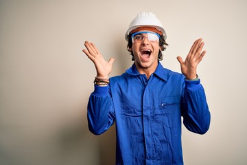 Young constructor man wearing uniform and security helmet over isolated white background celebrating crazy and amazed for success with arms raised and open eyes screaming excited. Winner concept