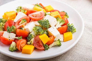 Vegetarian salad with broccoli, tomatoes, feta cheese, and pumpkin on white ceramic plate on a gray concrete background, side view, close up.