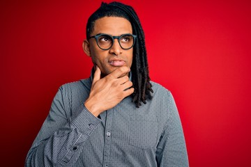 Young handsome african american man with dreadlocks wearing casual shirt and glasses with hand on chin thinking about question, pensive expression. Smiling with thoughtful face. Doubt concept.