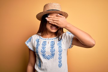 Young beautiful asian girl wearing casual t-shirt and hat standing over yellow background smiling and laughing with hand on face covering eyes for surprise. Blind concept.