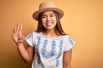 Young beautiful asian girl wearing casual t-shirt and hat standing over yellow background showing and pointing up with fingers number four while smiling confident and happy.