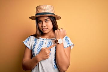 Young beautiful asian girl wearing casual t-shirt and hat standing over yellow background In hurry...