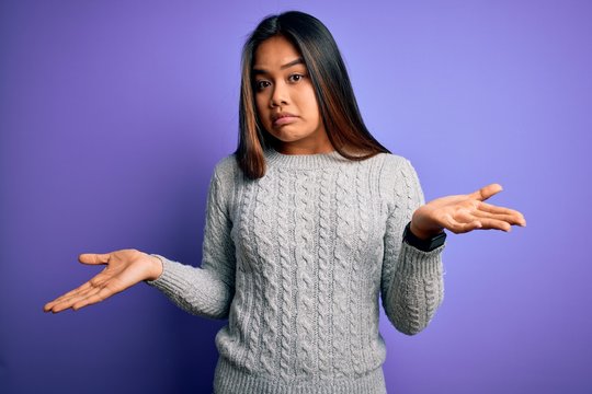 Young beautiful asian girl wearing casual sweater standing over isolated purple background clueless and confused expression with arms and hands raised. Doubt concept.