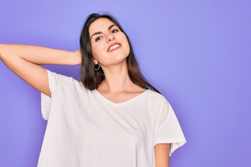 Young beautiful brunette woman wearing casual white t-shirt over purple background smiling confident touching hair with hand up gesture, posing attractive and fashionable