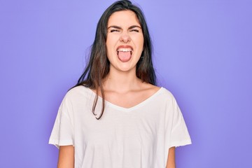 Fototapeta premium Young beautiful brunette woman wearing casual white t-shirt over purple background sticking tongue out happy with funny expression. Emotion concept.