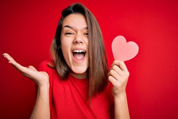 Young beautiful brunette romantic girl holding red paper heart shape over isolated background very happy and excited, winner expression celebrating victory screaming with big smile and raised hands