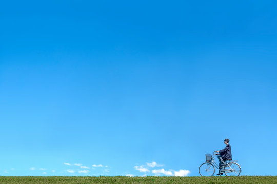 青空背景で緑の土手を自転車に乗る男子高校生。進学,入学,進路,通学イメージ