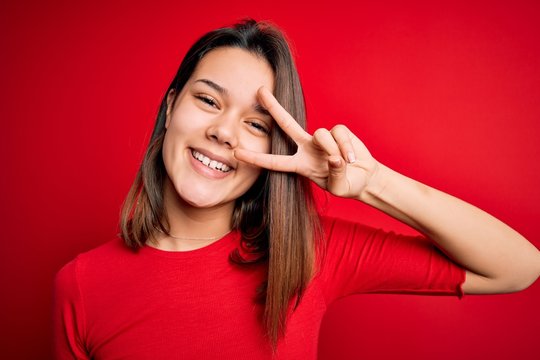 Young beautiful brunette girl wearing casual t-shirt over isolated red background Doing peace symbol with fingers over face, smiling cheerful showing victory