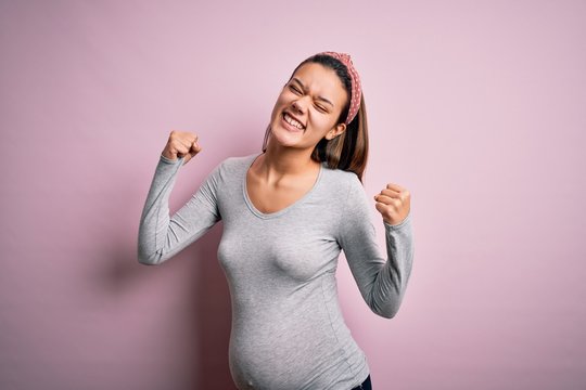 Young beautiful teenager girl pregnant expecting baby over isolated pink background very happy and excited doing winner gesture with arms raised, smiling and screaming for success. Celebration conc