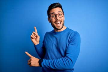 Young handsome man with beard wearing casual sweater and glasses over blue background smiling and...