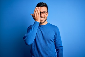 Young handsome man with beard wearing casual sweater and glasses over blue background covering one eye with hand, confident smile on face and surprise emotion.