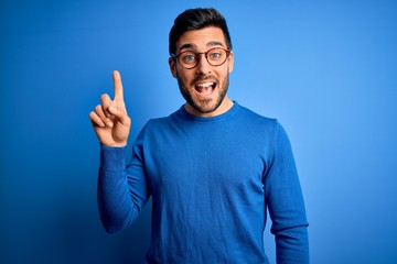 Young handsome man with beard wearing casual sweater and glasses over blue background pointing...