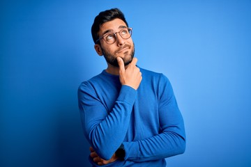 Young handsome man with beard wearing casual sweater and glasses over blue background Thinking...