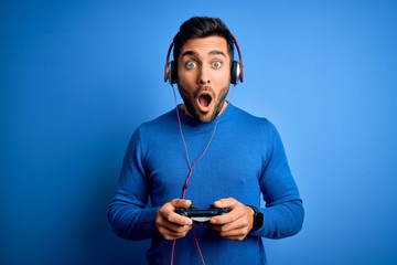 Young handsome gamer man with beard playing video game using joystick and headphones scared in shock with a surprise face, afraid and excited with fear expression © Krakenimages.com
