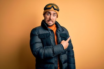 Young handsome skier man with beard wearing snow sportswear and ski goggles Pointing aside worried and nervous with forefinger, concerned and surprised expression