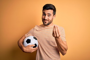 Handsome player man with beard playing soccer holding footballl ball over yellow background doing...
