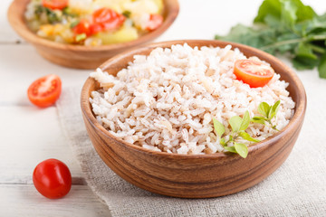 Unpolished rice porridge with stewed vegetables and oregano in wooden bowl on a white wooden background. side view, close up.