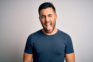 Young handsome man wearing casual t-shirt standing over isolated white background sticking tongue...
