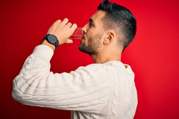 Young handsome man drinking glass of healthy water to refreshment standing over isolated red background