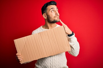 Young handsome man holding blank cardboard banner over isolated red background serious face...