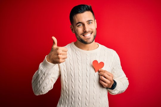 Young handsome man holding red shape heart as romantic and health symbol happy with big smile doing ok sign, thumb up with fingers, excellent sign