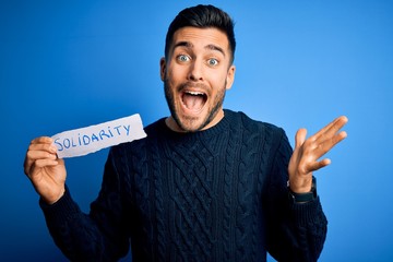 Young handsome man holding paper with solidarity message over blue background very happy and...
