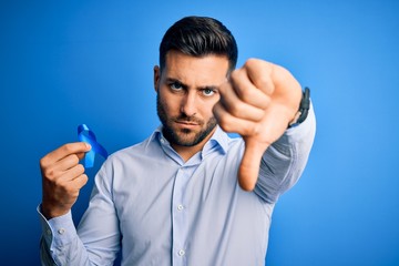 Young handsome man holding blue ribbon as prostate campaing support over blue background with angry face, negative sign showing dislike with thumbs down, rejection concept