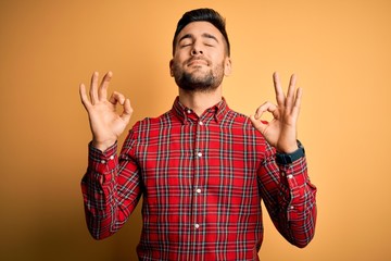 Naklejka premium Young handsome man wearing casual shirt standing over isolated yellow background relax and smiling with eyes closed doing meditation gesture with fingers. Yoga concept.