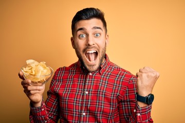 Young handsome man holding bowl with potato chips over isolated yellow background serious face thinking about question, very confused idea