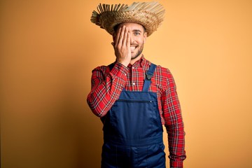 Obraz premium Young rural farmer man wearing bib overall and countryside hat over yellow background covering one eye with hand, confident smile on face and surprise emotion.
