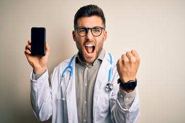 Young doctor man wearing stethoscope showing smartphone screen over isolated background annoyed and frustrated shouting with anger, crazy and yelling with raised hand, anger concept