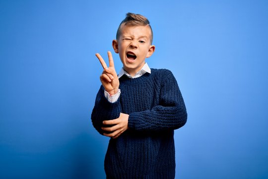 Young little caucasian kid with blue eyes wearing winter sweater over blue background smiling with happy face winking at the camera doing victory sign. Number two.