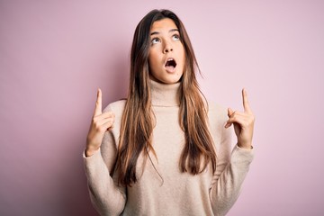 Young beautiful girl wearing casual turtleneck sweater standing over isolated pink background amazed and surprised looking up and pointing with fingers and raised arms.
