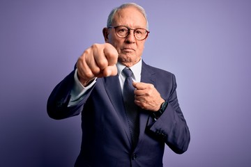 Grey haired senior business man wearing glasses and elegant suit and tie over purple background Punching fist to fight, aggressive and angry attack, threat and violence