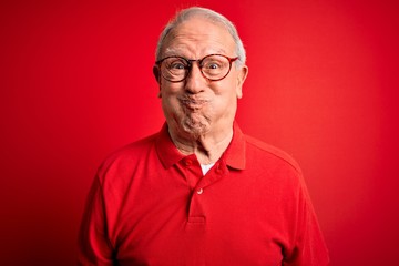 Grey haired senior man wearing glasses and casual t-shirt over red background puffing cheeks with funny face. Mouth inflated with air, crazy expression.