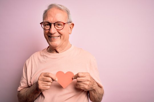 Senior grey haired man holding heart shape paper over pink background with a happy face standing and smiling with a confident smile showing teeth