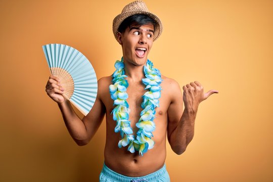Young tourist man on vacation wearing swimwear and hawaiian lei flowers holding hand fan pointing and showing with thumb up to the side with happy face smiling