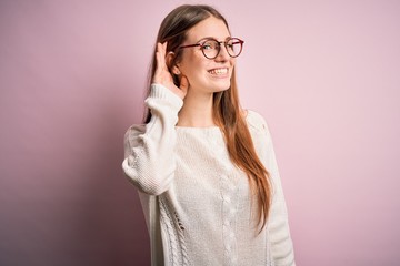 Young beautiful redhead woman wearing casual sweater and glasses over pink background smiling with hand over ear listening an hearing to rumor or gossip. Deafness concept.
