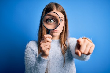 Young beautiful redhead detective woman using magnifying glass over isolated blue background pointing with finger to the camera and to you, hand sign, positive and confident gesture from the front