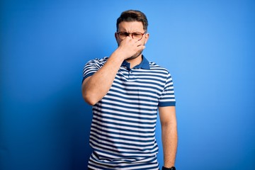Young man with blue eyes wearing glasses and casual striped t-shirt over blue background smelling...