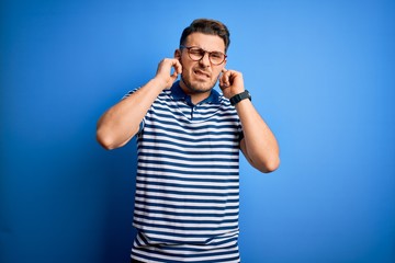 Fototapeta premium Young man with blue eyes wearing glasses and casual striped t-shirt over blue background covering ears with fingers with annoyed expression for the noise of loud music. Deaf concept.