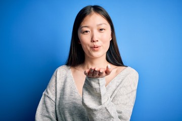 Obraz premium Young beautiful asian woman wearing casual sweater standing over blue isolated background looking at the camera blowing a kiss with hand on air being lovely and sexy. Love expression.
