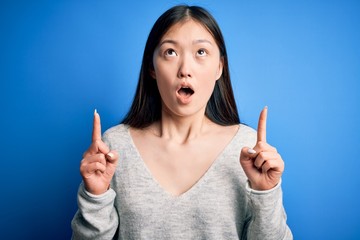 Young beautiful asian woman wearing casual sweater standing over blue isolated background amazed and surprised looking up and pointing with fingers and raised arms.