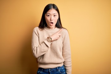 Young beautiful asian woman wearing casual sweater over yellow isolated background Surprised pointing with finger to the side, open mouth amazed expression.