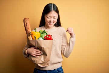 Young asian woman holding paper bag of fresh healthy groceries over yellow isolated background very happy and excited doing winner gesture with arms raised, smiling and screaming for success